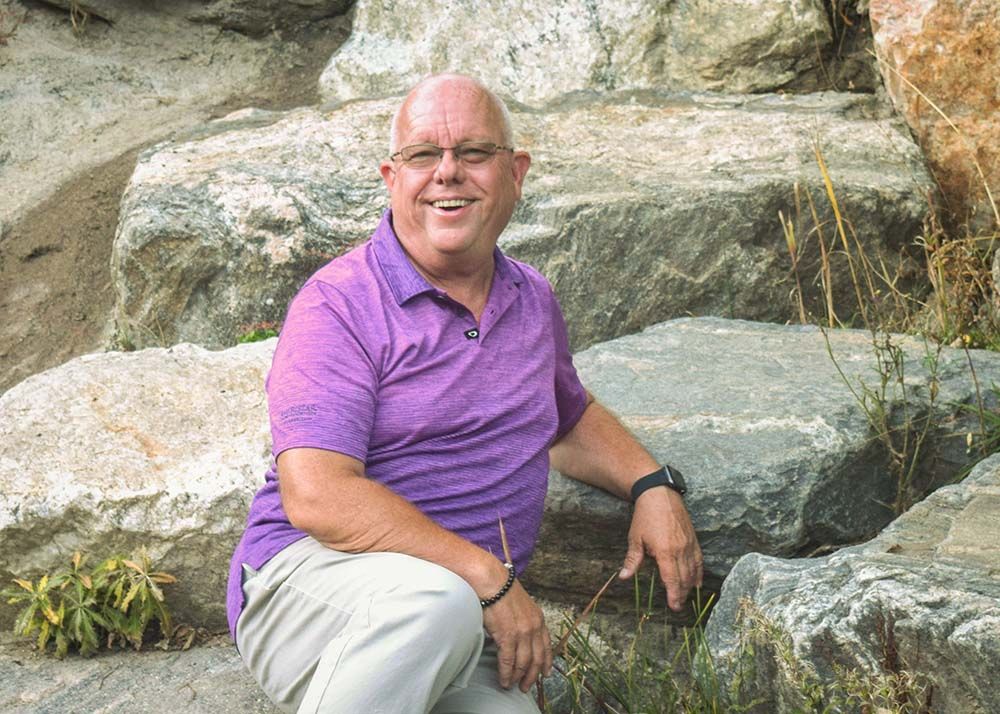 Kevan smiling in a purple polo casually leaning on boulders near a river