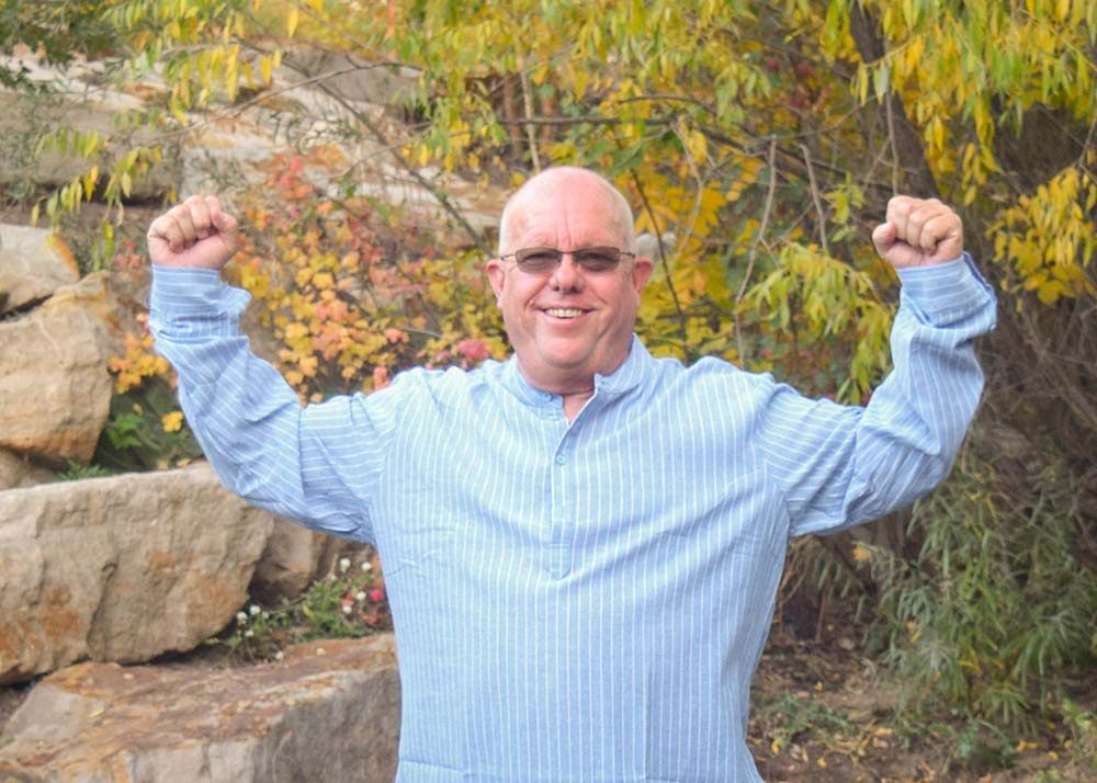 Kevan smiling in a blue long sleeve shirt flexing in nature